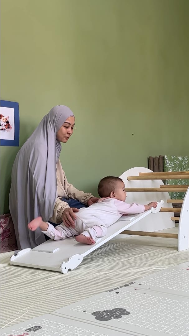 Pikler qiyaligida chaqaloqni qo'llab turgan ota-ona
